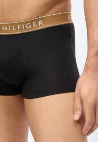 Άνδρας που φοράει μαύρα μποξεράκια με καφέ ζώνη μέσης που φέρει την επωνυμία «HILFIGER», το χέρι να στηρίζεται στο πλάι του.