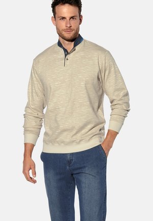 Beige strukturierter Pullover mit einem marineblauen Kragen und drei Knöpfen, kombiniert mit blauen Jeans. Die Ärmel sind am Bund gerippt.