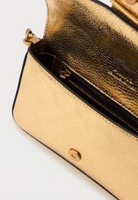Bolsa com textura dourada e borda preta. Apresenta um bolso interior com fecho e um fecho metálico circular. O interior tem um revestimento brilhante.