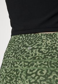Leggings atléticos com padrão verde e material elástico, apresentando um discreto logotipo prateado e uma costura plana para conforto. Um top preto é visível por cima.