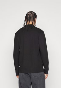 Person med flätat hår som bär en svart långärmad tröja och grå jeans, st ående vänd bort mot en enkel ljus bakgrund.