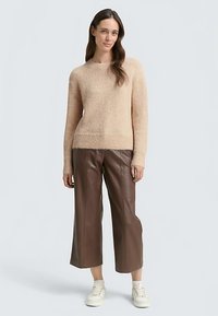 Beiger Strickpullover mit Rundhalsausschnitt und geripptem Saum, kombiniert mit braunen, kurzen Lederhosen und weißen Sneakers. Glatte Textur, lässige Passform.