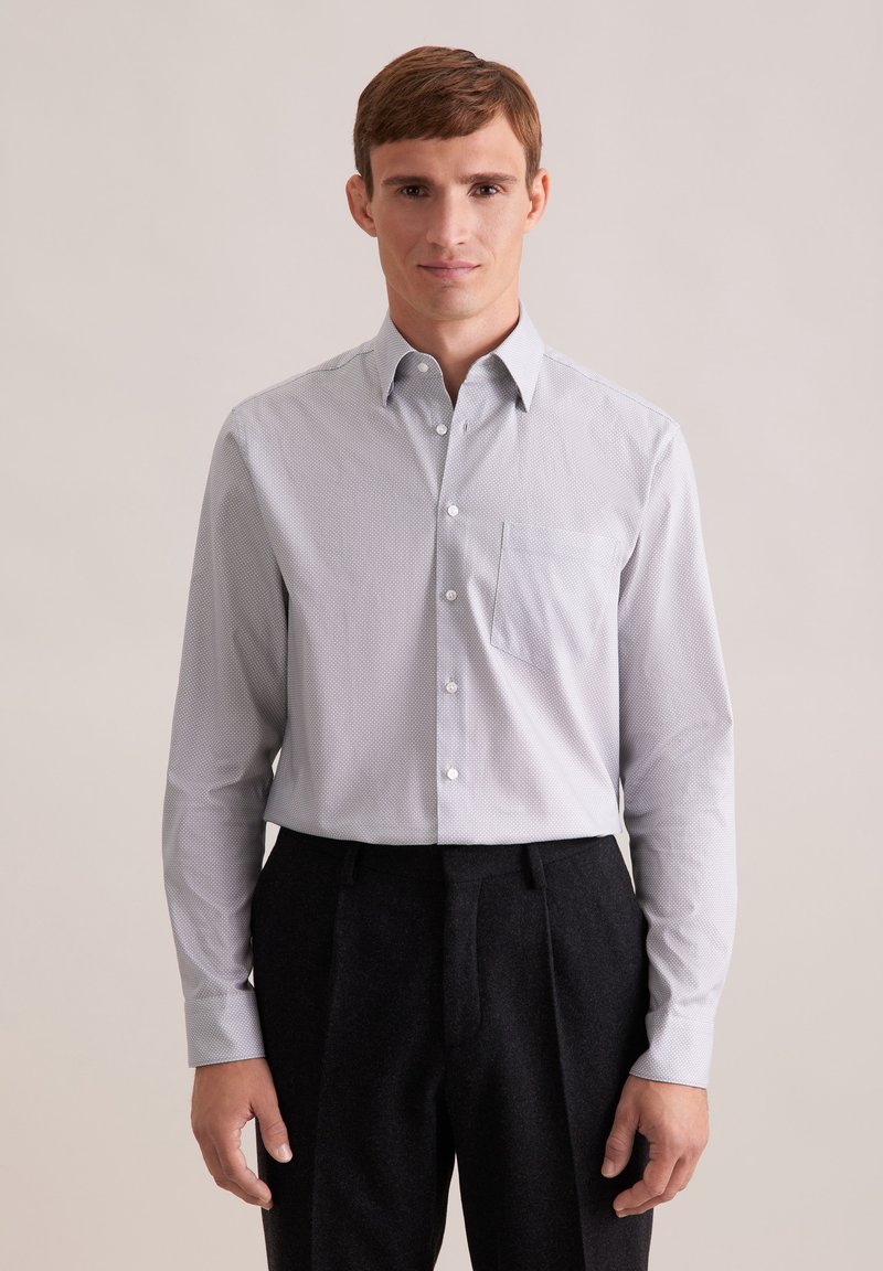 Camicia grigia chiaro con una piccola trama geometrica, taschino sul petto singolo e maniche lunghe, abbinata a pantaloni sartoriali scuri.