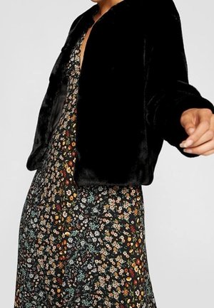 Osoba ubrana w czarną kurtkę z futerka ekologicznego na długą czarną sukienkę z małym, wielokolorowym wzorem kwiatowym, sięgającą do połowy łydki.