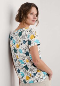 Korte mouw shirt met een witte achtergrond en een bloemenpatroon in teal, geel en grijs. Zachte stof en ontspannen pasvorm ontwerp.