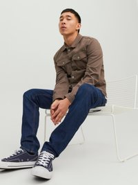 Brun bomullsjakke med store klaffelommer, knappdetaljer og krage, kombinert med mørkeblå jeans og marineblå sneakers. Sittende på en hvit stol.
