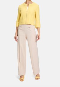 Žlutá zkrácená bunda s kulatým límcem a předním zipem, kombinována s béžovými kalhotami s širokými nohavicemi a zlatými knoflíky a žlutými podpatky.