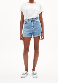 Pantalones cortos de mezclilla con un lavado en azul claro, de cintura alta y con puños enrollados, combinados con una camiseta blanca de manga corta y zapatillas con acentos azules.