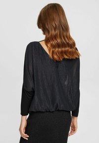 Top lungo nero a maniche con scollatura a V profonda sul retro, realizzato in tessuto testurizzato con un leggero luccichio. Presenta una vita arricciata e maniche aderenti.