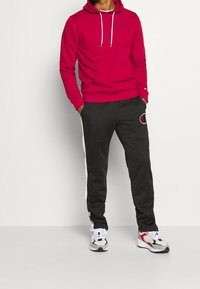 Felpa rossa in cotone con tasca frontale e lacci bianchi, abbinata a pantaloni jogger neri con una striscia laterale e dettaglio del logo.