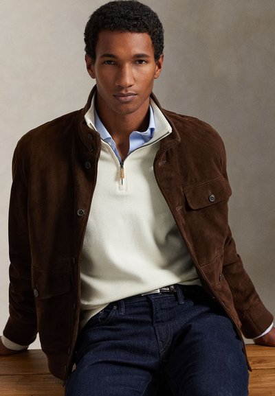 Νέος άνδρας που φορά καφέ σουέντ σακάκι πάνω από ένα κρεμ πουλόβερ με μισό φερμουάρ και μπλε πουκάμισο, καθισμένος σε ξύλινη επιφάνεια με ουδέτερο φόντο.