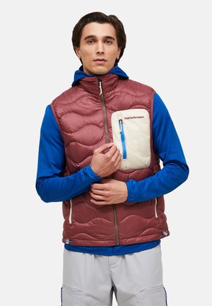 Mand iført bordeaux quiltet vest med cremefarvet brystlomme over blå hoodie, der justerer vestens lynlås, stående foran hvid baggrund.