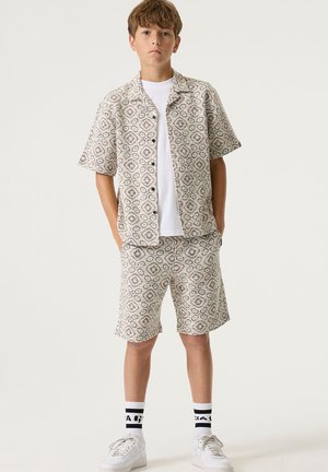 Korte mouwen, blouse met knopen en bijpassende korte broek, met een patroon in beige, roze en grijs. Draagt een wit T-shirt en witte sneakers.