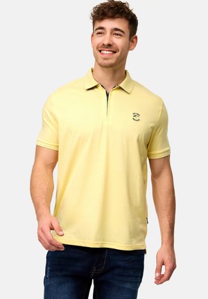 Gul polo-shirt med korte ærmer, krave og frontlynlås. Har et lille logo i marineblå på venstre bryst. Glat bomuldsstof.