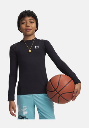 Chlapec v černém tričku s dlouhým rukávem a modrých šortkách drží basketbal s pravou rukou na bílém pozadí.