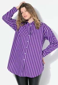 Camicia viola a maniche lunghe con righe bianche, apertura con bottoni frontale, taschino a sinistra e vestibilità comoda. Abbinata a leggings neri.