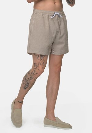 Man draagt beige gestreepte shorts en beige instapschoenen, toont beentatoeages waaronder gezicht- en bloemenmotieven.
