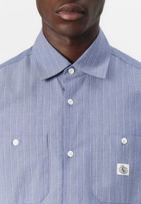 Blåstripete skjorte med knapper laget av lett stoff, med to frontlommer og hvite knapper. Inkluderer et brodert logo på lommen.