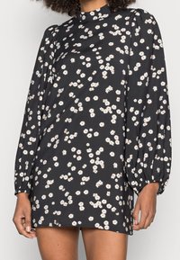 Schwarzes Kleid mit weißem Gänseblümchenmuster; hoher Kragen; lange, puffige Ärmel; weicher Stoff; entspannte Passform; knielange Länge.