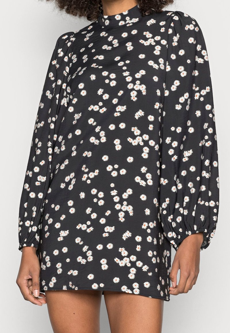 Schwarzes Kleid mit weißem Gänseblümchenmuster; hoher Kragen; lange, puffige Ärmel; weicher Stoff; entspannte Passform; knielange Länge.