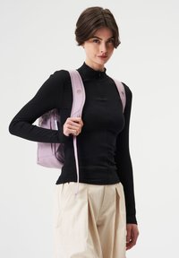 Schwarzes, geripptes Langarmoberteil kombiniert mit cremefarbenen, weiten Hosen; das Model trägt einen weichen, lila Rucksack mit schmalen Trägern und runden Verschlüssen.