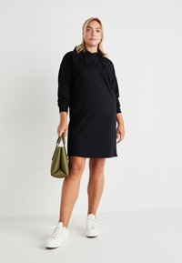 Črna obleka s kapuco, z teksturiranimi čipkastimi poudarki na ramenskem delu. Obuta v bele superge in drži zeleno torbico.