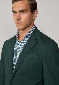 Blazer vert foncé en coton avec un revers cranté, une poche poitrine simple et une fermeture boutonnée, associé à une chemise à col rayée.