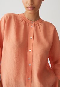 Camicia button-up corallo a vestibilità morbida con scollatura rotonda, texture sottile e bottoni bianchi sul davanti.