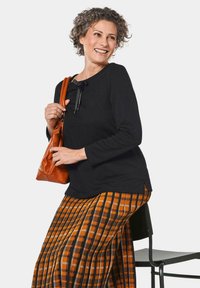 Schwarzes Langarmtop mit rundem Ausschnitt und einem Schleifendetail, kombiniert mit einem karierten orange-schwarzen Faltenrock. Braune Handtasche.