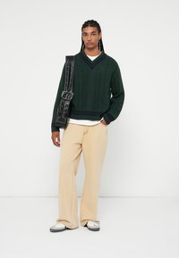 Mörkgrön stickad tröja med V-hals, tillsammans med krämfärgade vida byxor och en svart axelväska. Sneakers fulländar outfiten.