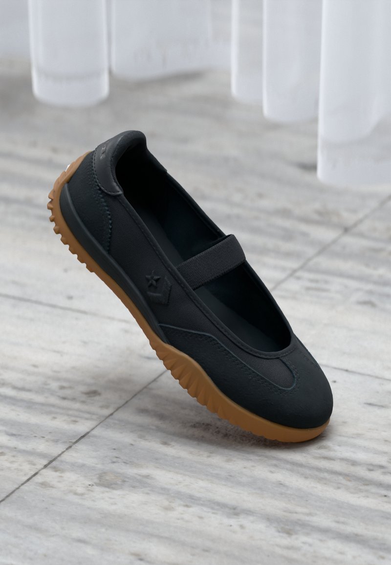 Chaussure noire style Mary Jane avec bride élastique et semelle texturée marron, posée sur un sol en marbre avec un fond de rideau blanc.