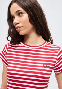 Νεαρή γυναίκα με μακριά καστανά μαλλιά, φορώντας κόκκινη και λευκή ριγέ μπλούζα με τον λογότυπο Pepe Jeans, κοιτάζοντας ελαφρώς προς τα αριστερά.