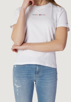 Vrouw draagt een witte Tommy Jeans T-shirt met een meerkleurig logo en lichtblauwe distressed jeans, armen gekruist met hand die het gezicht raakt.