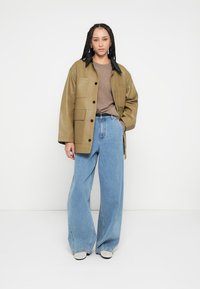 Brūnganas ādas jaka ar melnu apkakli, lielām priekšējām kabatām, valkāta kopā ar gaiši zilām, platiem džinsiem un baltām kurpēm.