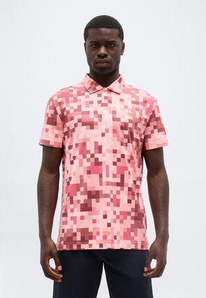 Mężczyzna ubrany w różowo-czerwoną, krótki rękaw, koszulkę polo z pikselowym wzorem, stojący na tle jasnej, jednolitej ściany.