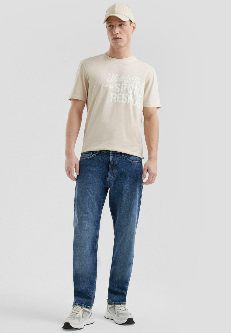 T-shirt in cotone beige con testo grafico, jeans in denim blu e scarpe da ginnastica grigie. Un cappellino beige chiaro completa l'outfit.