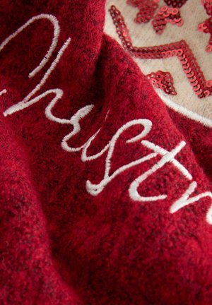 Roter Textilstoff mit weißer, geschwungener Stickerei und einem beigen Patch, der mit roten Pailletten im Sternen-Muster verziert ist.