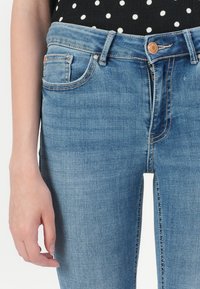 Person som har på seg lyseblå denimbukser med delvis åpen glidelås og en svart topp med hvite prikker synlig ved midjen.