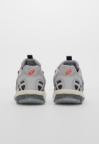 Harmaat urheilujalkineet, joissa on teksturoitu verkko-yläosa, mustat yksityiskohdat ja valkoinen välipohja. Takana on pieni musta vetotappi ja logo-yksityiskohta.