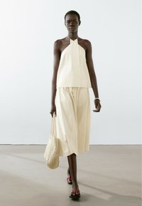 Crème-kleurige mouwloze top met halternek en textuurdesign, gecombineerd met wijde broek. Model draagt een geweven tas en heeft rode sandalen aan.
