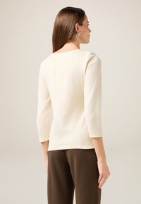 Crème driekwart mouwen top met een ronde halslijn, gladde textuur en een aansluitende silhouette, gecombineerd met een bruine broek.