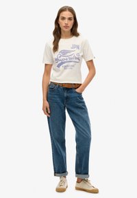 Hvid bomulds t-shirt med lilla grafisk tekst, parret med blå denim jeans. Jeansene er rullet op ved nedkanterne og båret med beige sneakers.