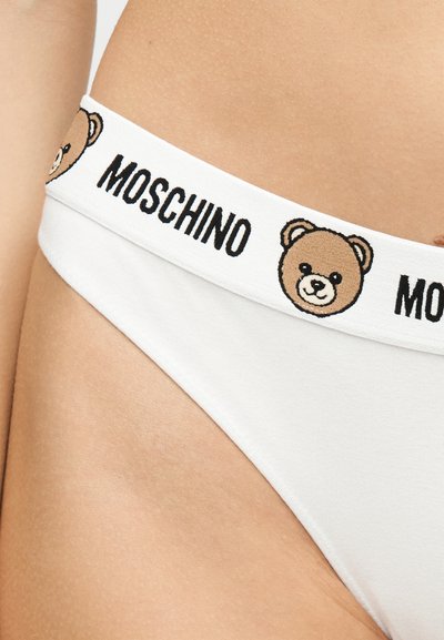 Λευκή μέση εσωρούχου με επαναλαμβανόμενα πρόσωπα αρκουδάκια και το λογότυπο "MOSCHINO", φορεμένη στη λεκάνη και τη μέση ενός ατόμου.