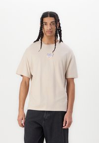Beige t-skjorte med korte ermer og rund hals, med en liten, reflekterende logo i midten. Glatt bomullsstoff med en avslappet passform.