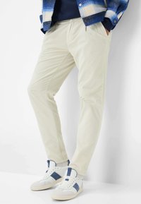 Pantaloni di velluto a coste crema con righe verticali, dotati di tasche laterali. Abbinati a sneakers bianche e blu, esposti su uno sfondo chiaro.