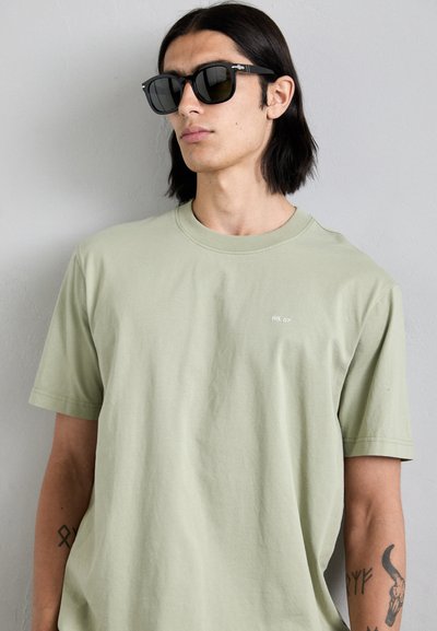 Tricou din bumbac de culoare verde deschis, cu gât rotund și mâneci scurte, având un mic logo alb. Ochelari de soare pătrați negri cu accente metalice.