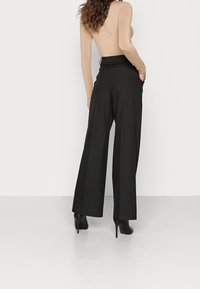 Kobieta z długimi kręconymi włosami, ubrana w beżową bluzkę z długim rękawem, czarne spodnie w szerokim kroju i czarne botki na wysokim obcasie, stoi pod białą ścianą.