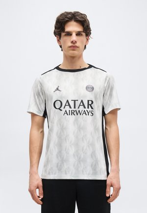 Fiatal férfi áll, fehér és fekete Paris Saint-Germain focimezt visel, amelyen a Qatar Airways logója látható.