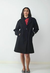 Schwarzer Wollmantel mit Reverskragen, knopfformiger Verschluss vorne und seitlichen Taschen. Glatte Textur und leicht ausgestellt am Saum. Modell trägt ein Oberteil mit rotem Akzent.