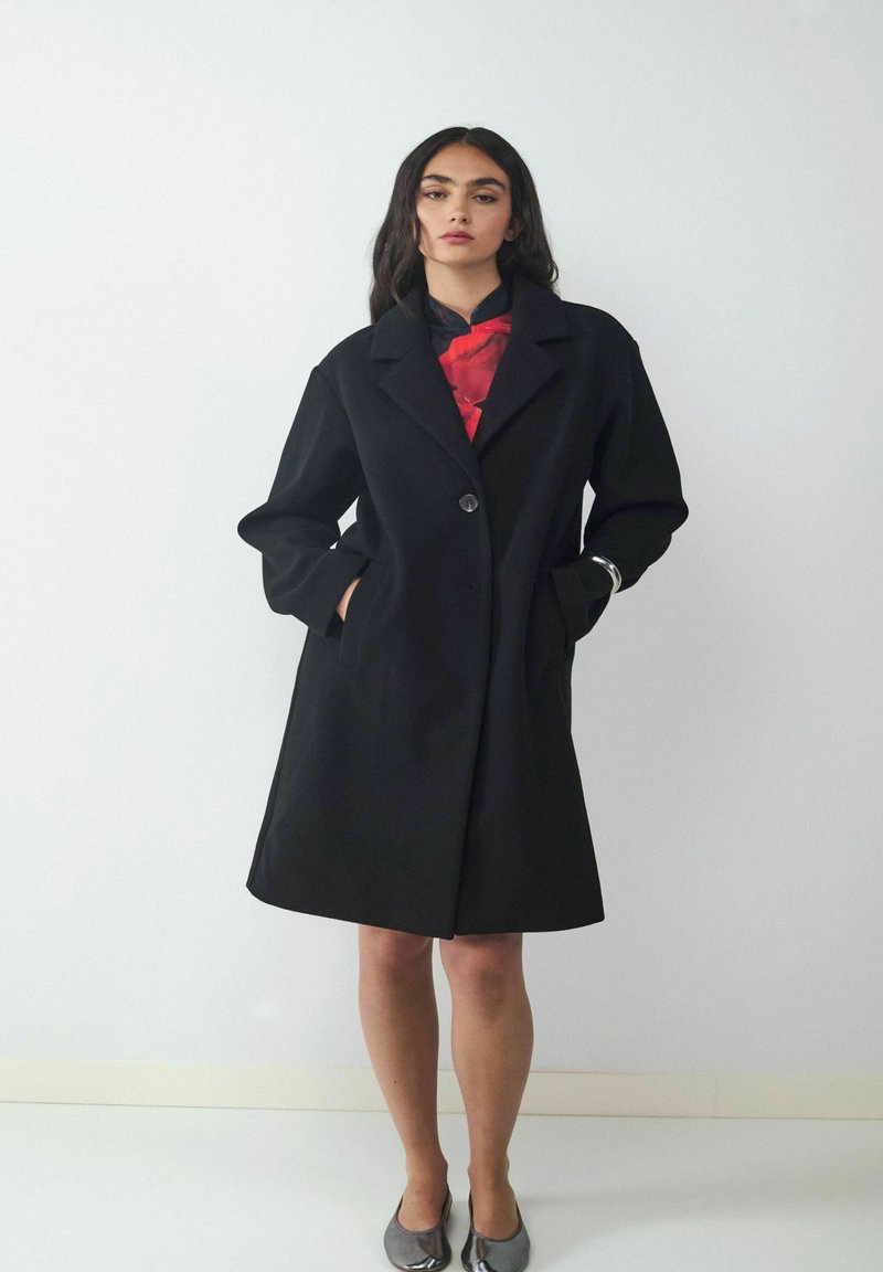 Schwarzer Wollmantel mit Reverskragen, knopfformiger Verschluss vorne und seitlichen Taschen. Glatte Textur und leicht ausgestellt am Saum. Modell trägt ein Oberteil mit rotem Akzent.